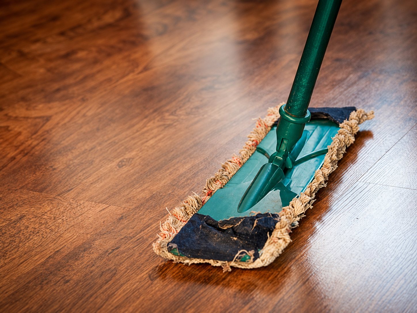 Un balai à franges vert glisse sur un parquet en bois foncé et brillant.