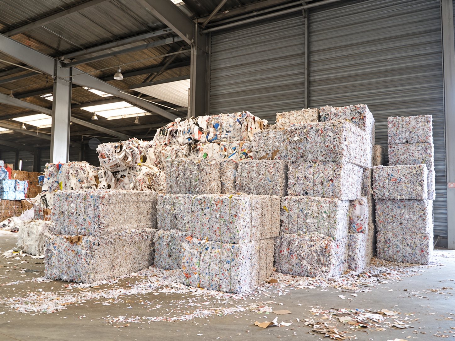 Des piles de grosses balles de papier et de carton compressés sont entreposées dans un grand hangar industriel, avec des déchets de papier au sol.