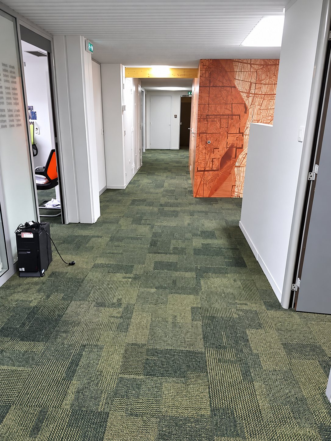 Un long couloir de bureaux au sol recouvert de moquette en dalles vertes, avec un mur décoré d'une carte orange et des portes vitrées sur le côté.