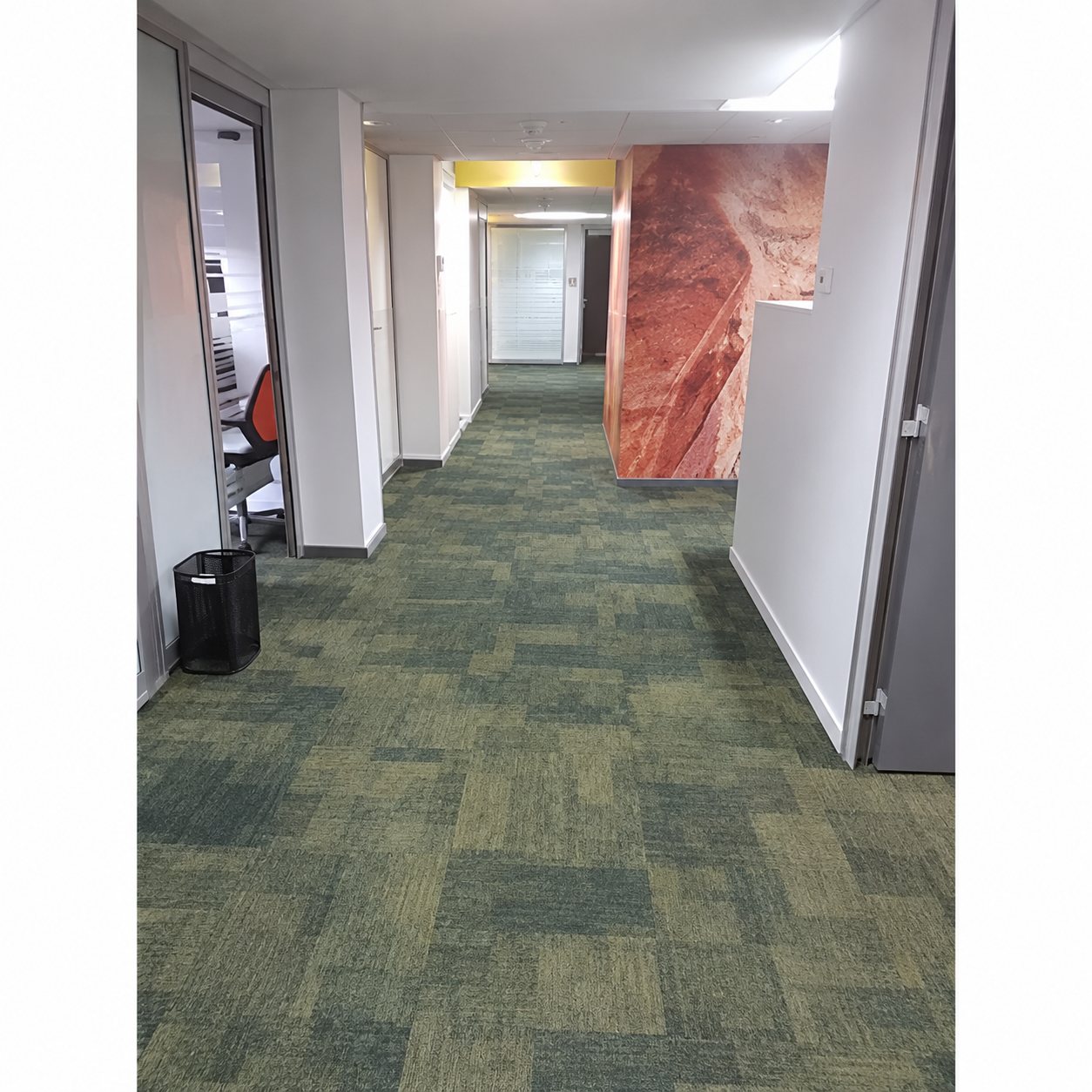 Un long couloir de bureaux au sol recouvert de moquette en dalles grises et vertes, avec des portes ouvertes sur les côtés et une paroi orangée au fond à droite.