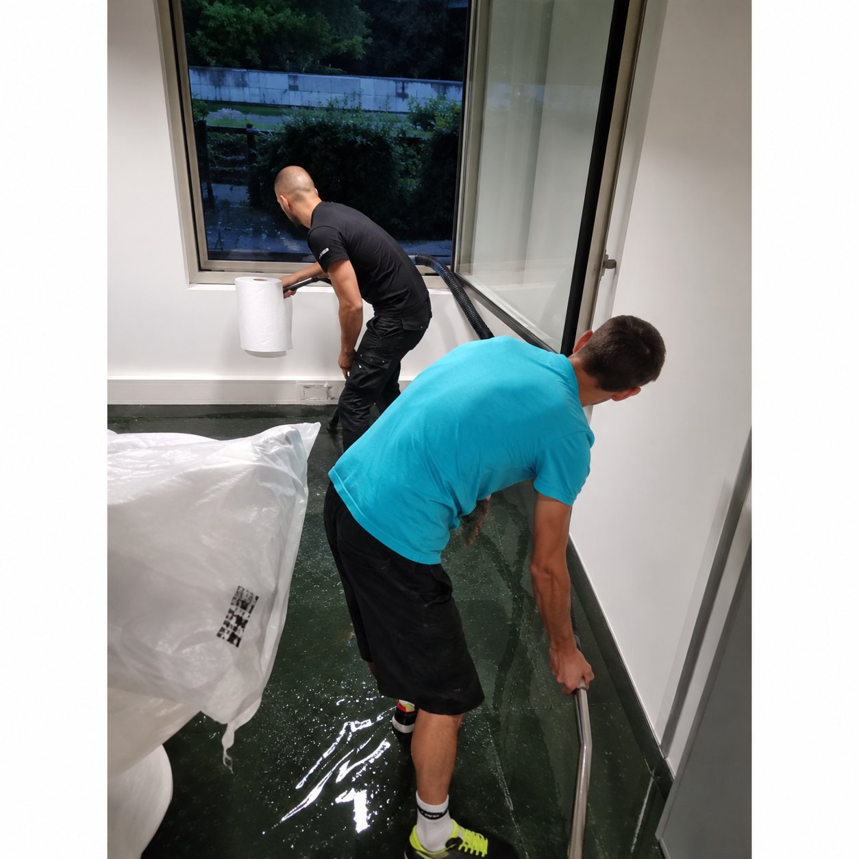 Deux hommes en tenue de travail nettoient l'intérieur d'une pièce lumineuse, l'un en t-shirt turquoise tenant un tuyau au premier plan.