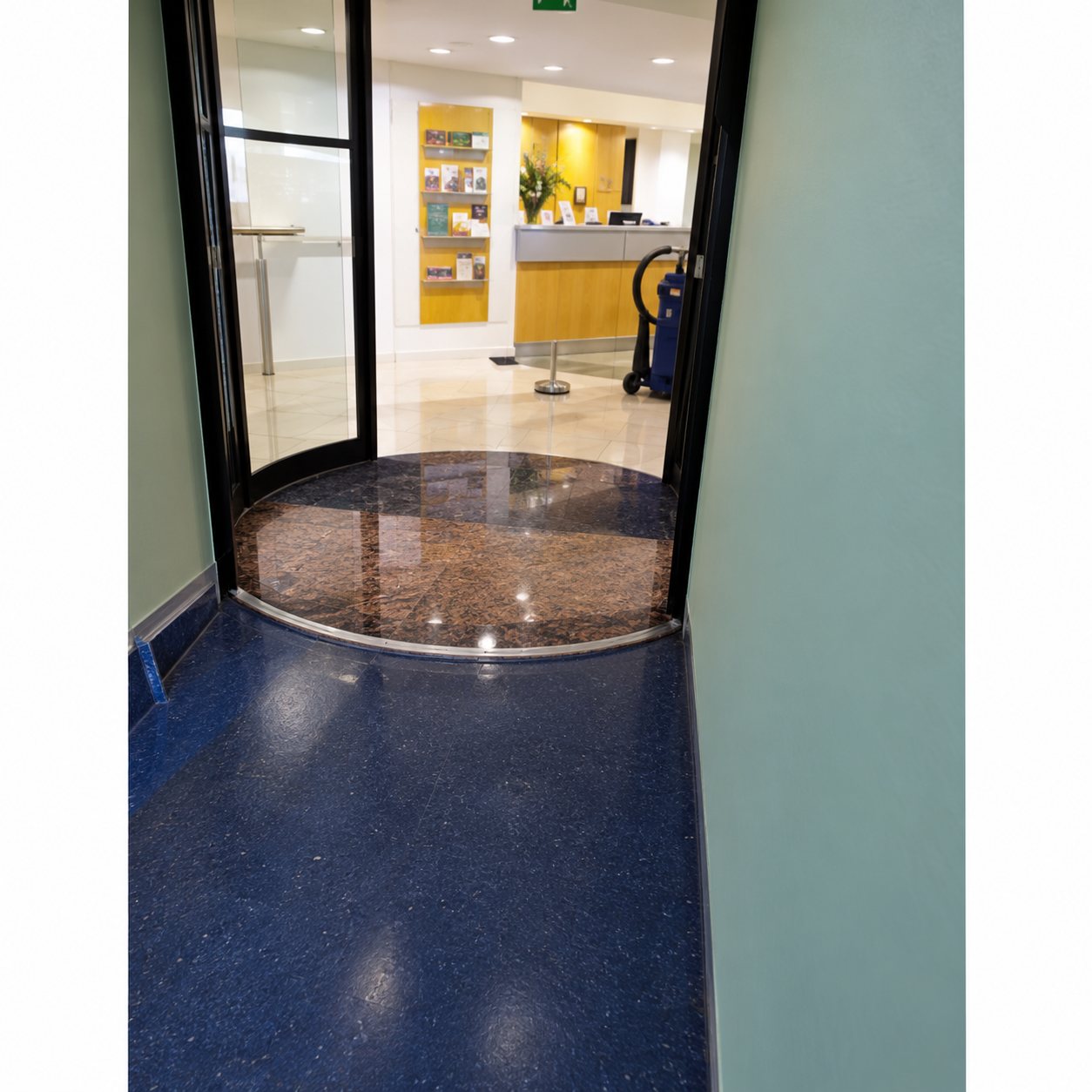 Vue d'un couloir au sol bleu menant à une porte tournante vitrée, avec un espace lumineux aux murs jaunes en arrière-plan.