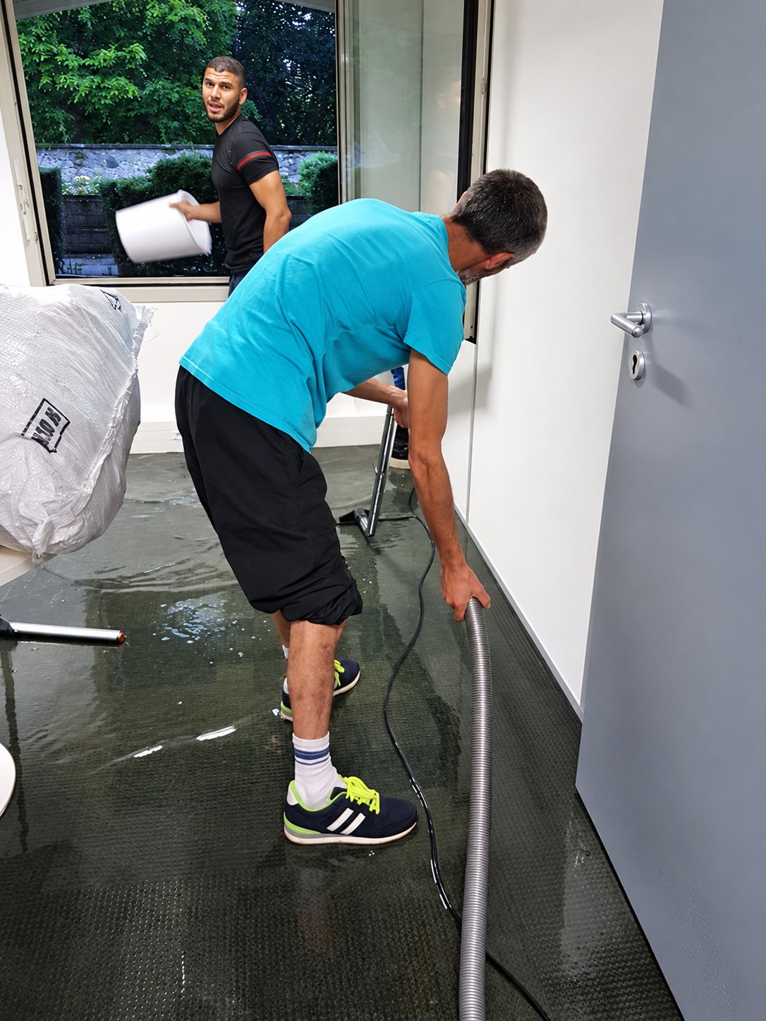 Deux hommes en t-shirt nettoient une pièce inondée, l'un avec un aspirateur à eau et l'autre portant un seau blanc près de la fenêtre.