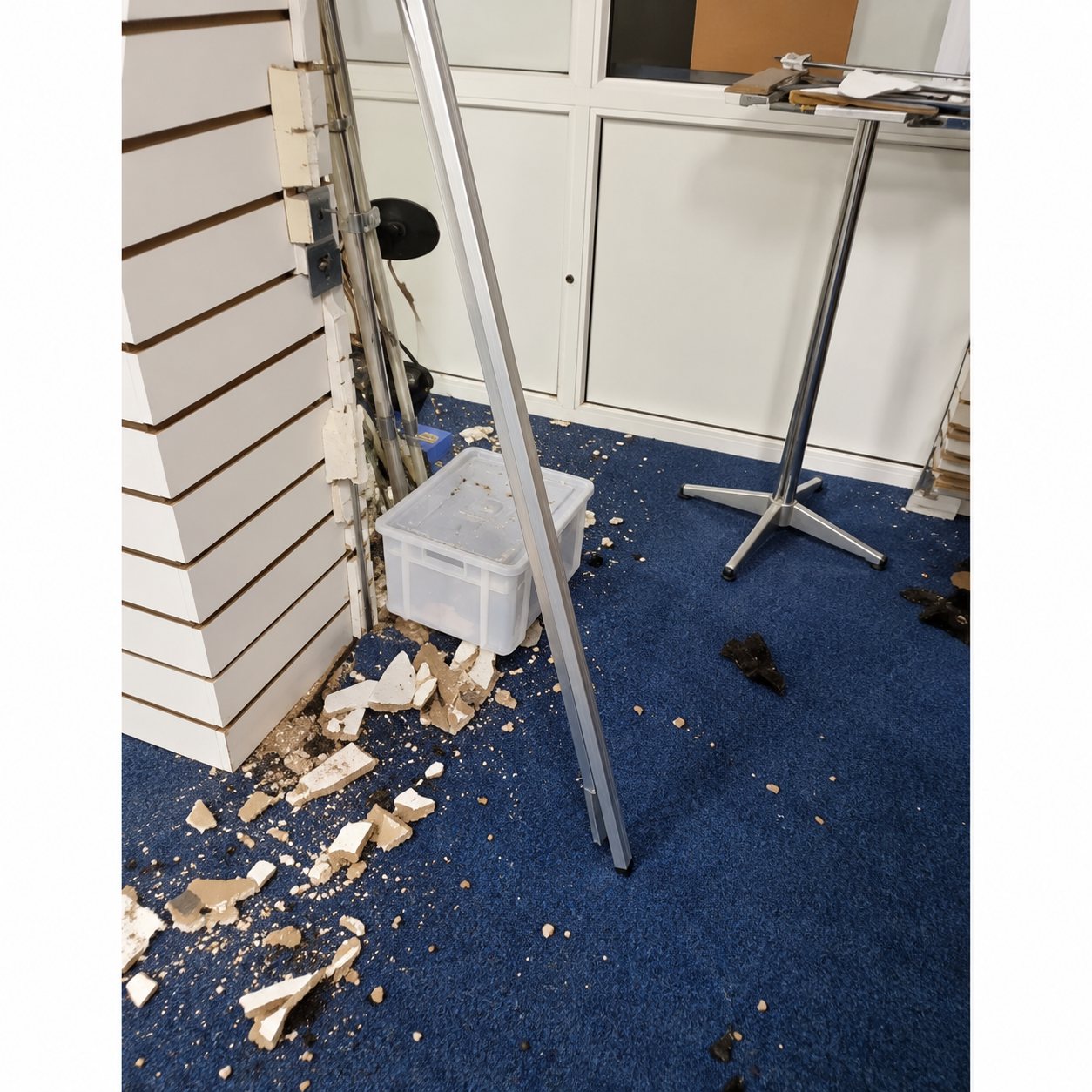 Intérieur d'un local commercial dont le sol bleu est jonché de débris, avec une étagère blanche à gauche et une table haute au fond.