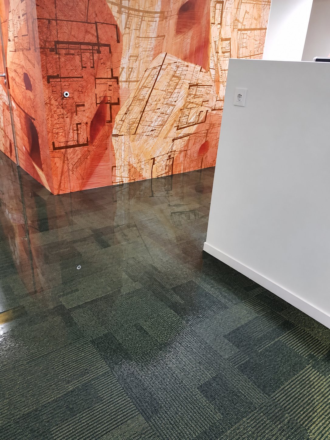 Une moquette de bureau gris-vert recouverte d'eau reflète un mur orange imprimé de plans d'architecte, à côté d'une cloison blanche.