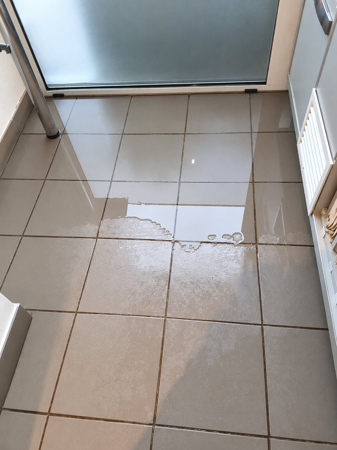 Une flaque d'eau recouvre le carrelage gris d'une pièce, devant une porte vitrée dépolie et une grille d'aération.