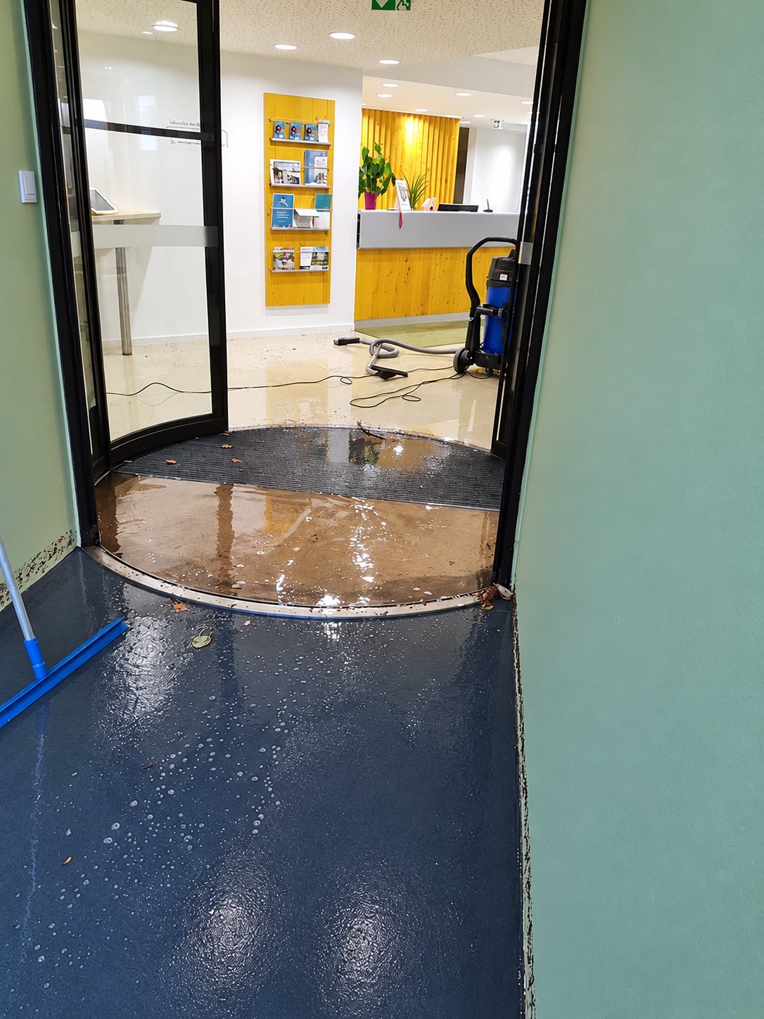 Une entrée de bâtiment avec porte tournante dont le sol est recouvert d'eau, tandis qu'un aspirateur eau et poussière est installé dans l'accueil au fond.