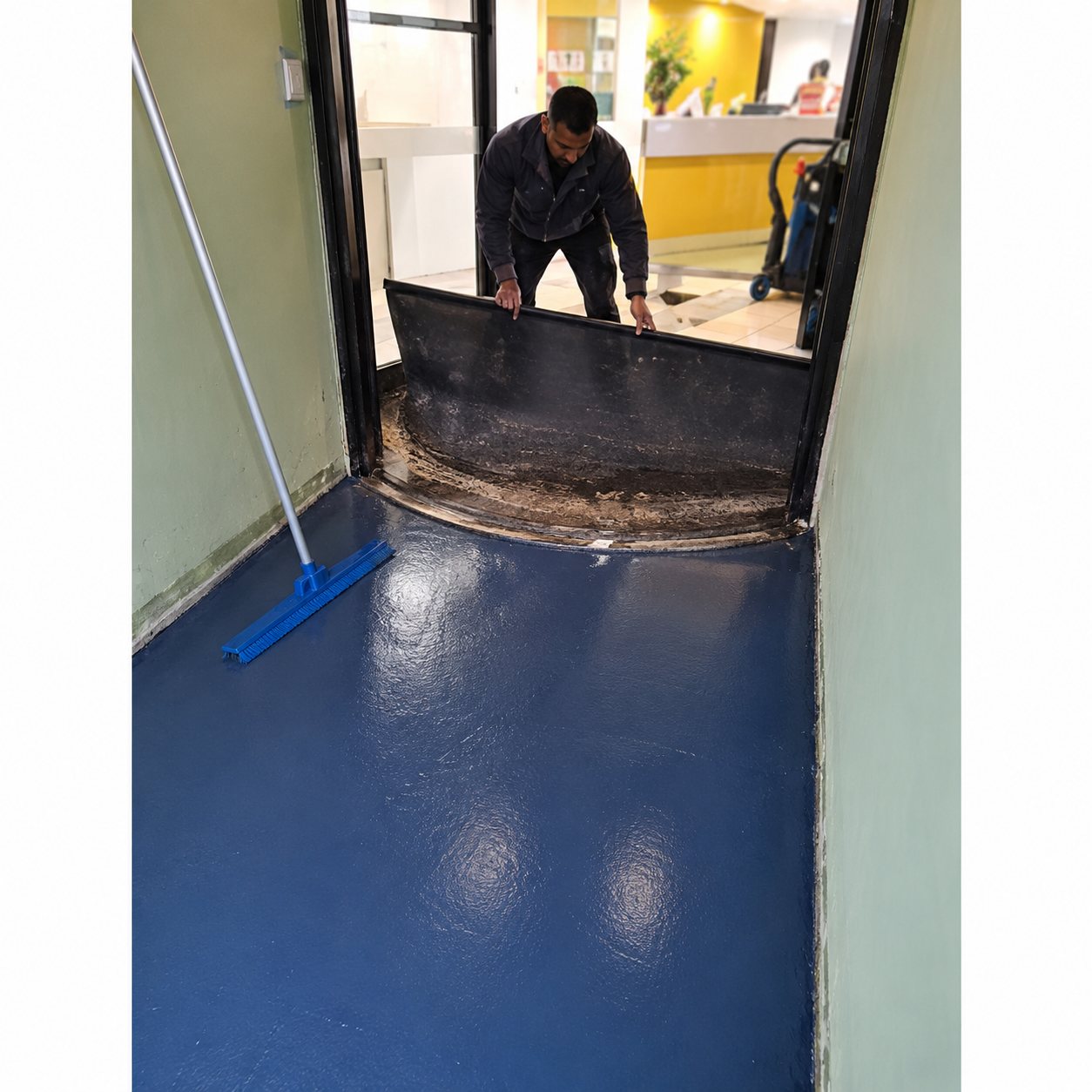 Un ouvrier travaille sur un sol fraîchement recouvert d'un revêtement bleu brillant, près d'une entrée vitrée.