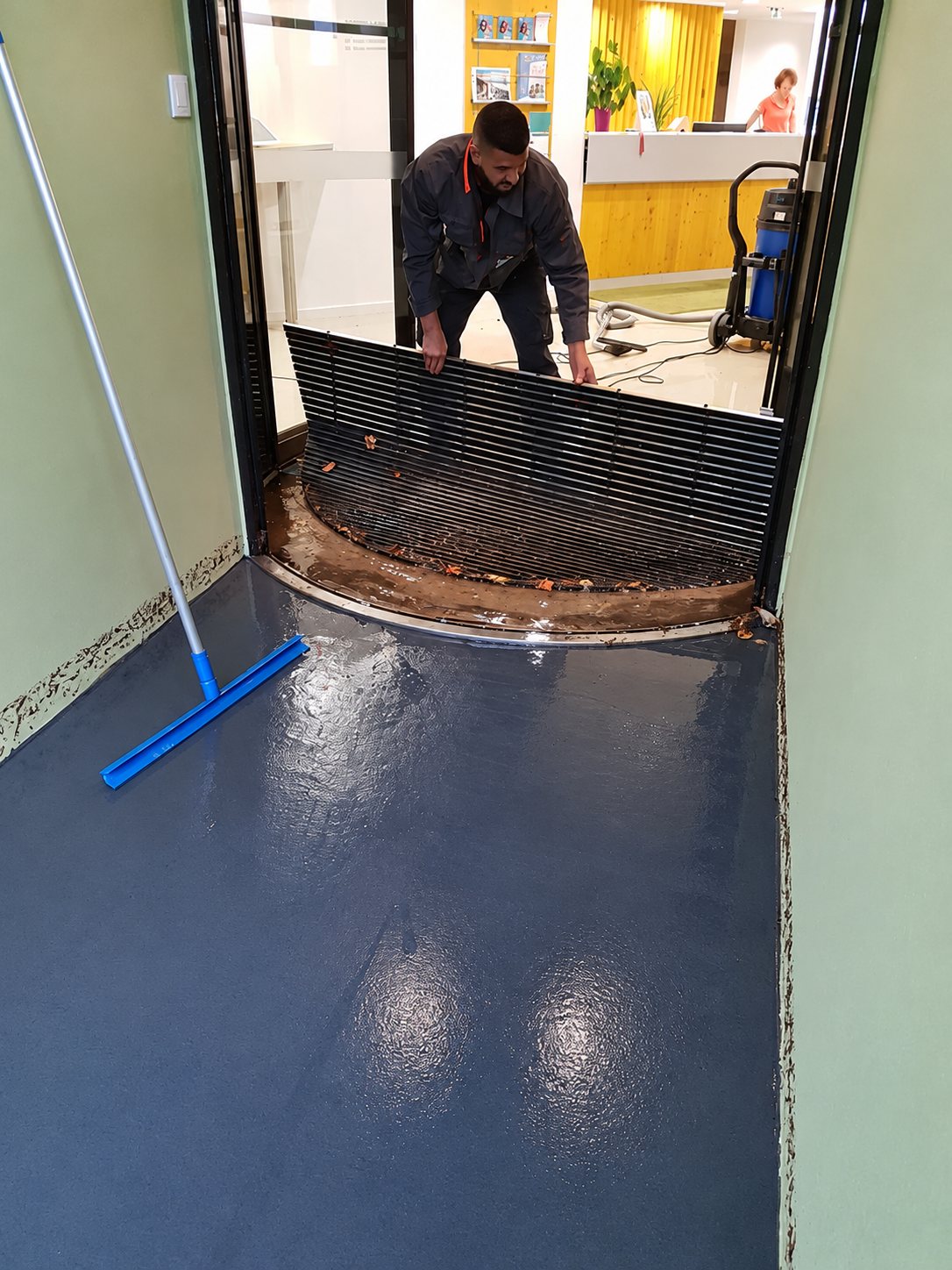 Un ouvrier soulève une grille de tapis d'entrée devant une porte vitrée, sur un sol fraîchement recouvert d'une résine bleu foncé brillante, avec une raclette et un aspirateur industriel à proximité.