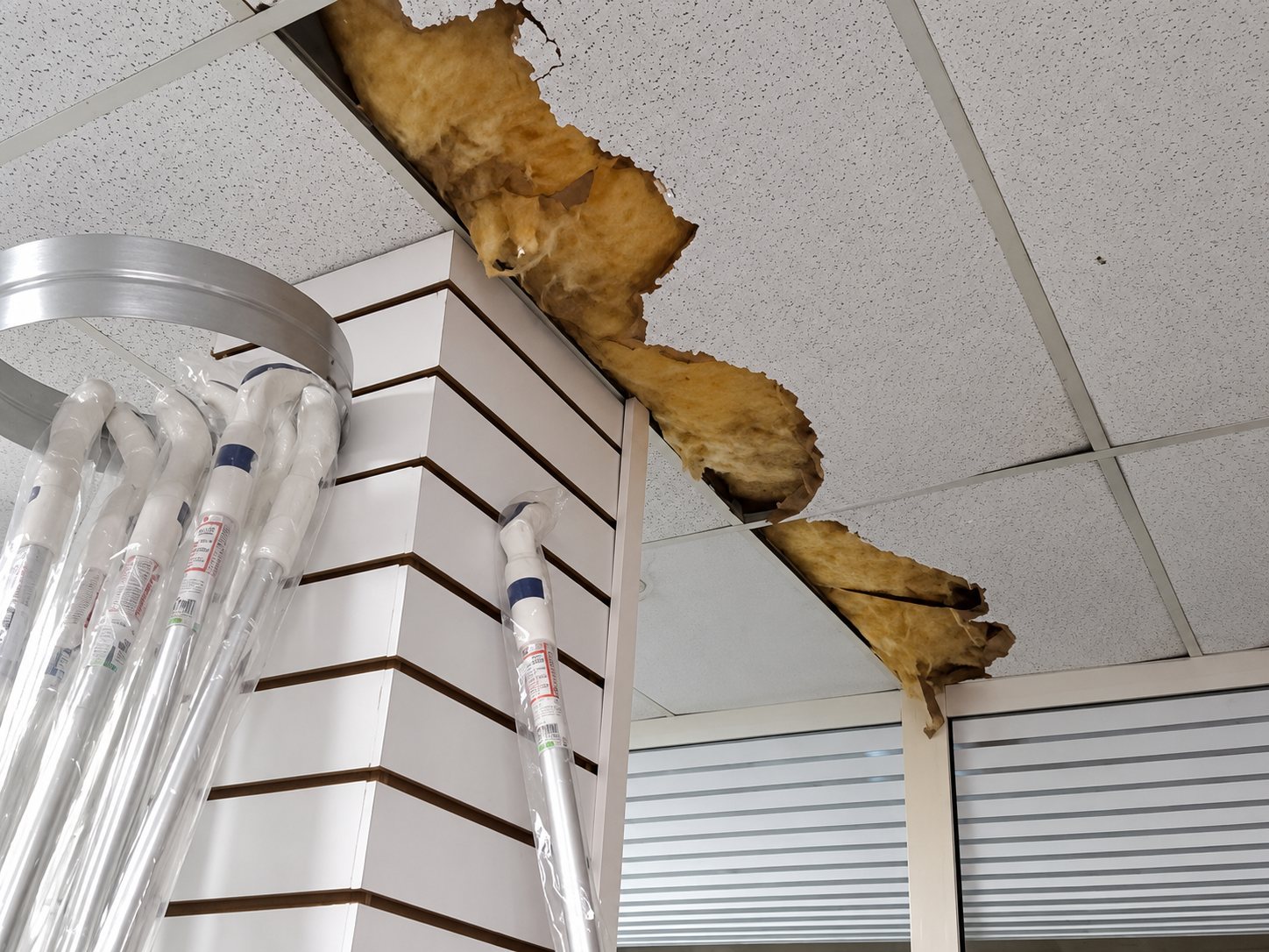 Dans un magasin, une dalle de plafond est endommagée et laisse apparaître de l'isolant, à côté d'un mur à lamelles et de tubes transparents exposés.