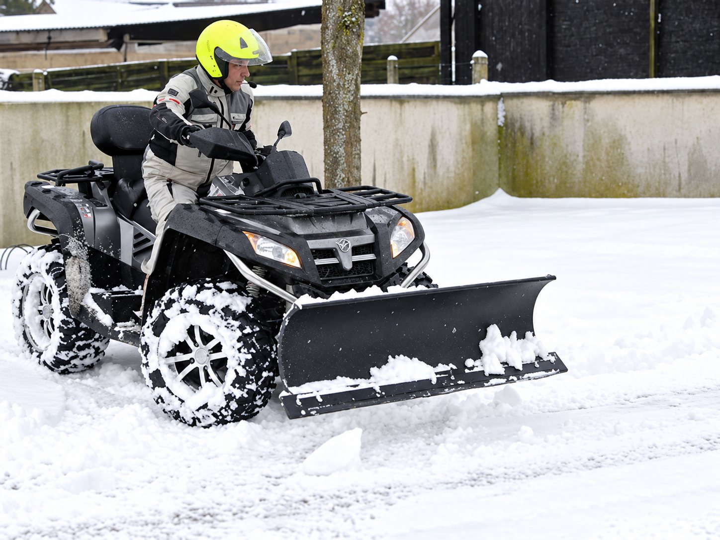 Une personne en casque jaune conduit un quad équipé d'une lame à neige et dégage une route couverte de neige devant un mur.