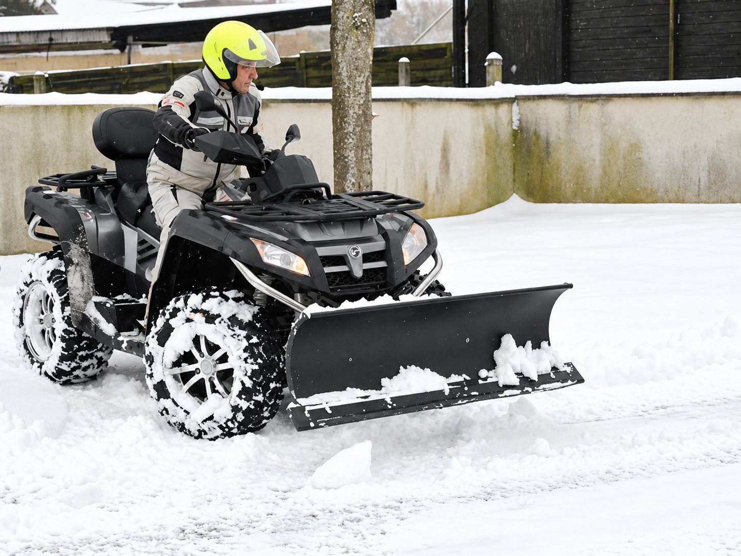 Une personne en casque jaune conduit un quad muni d'une lame à neige pour dégager une route recouverte de neige devant un mur.
