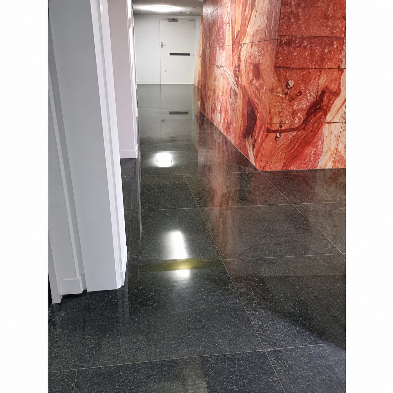 Un couloir intérieur dont le sol sombre et poli reflète la lumière, avec un mur rose texturé sur la droite.