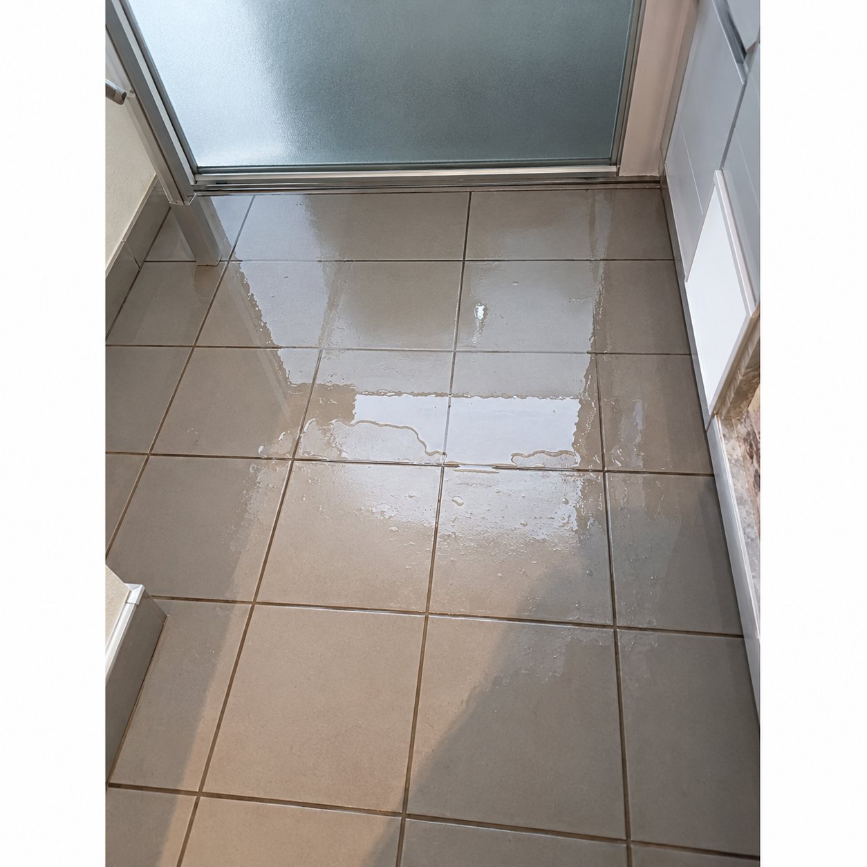 Sol de salle de bain recouvert de grands carreaux gris clair, avec une zone humide qui reflète la lumière, près d'une paroi de douche vitrée.